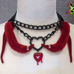 May include: A black choker necklace with two layers of black chain. Two red fuzzy worms with googly eyes are attached to the chain. A heart pendant with a dripping effect hangs from the center. The text "lordeplanec" is visible.