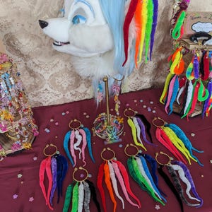 Puede incluir: Una exhibición de accesorios coloridos hechos a mano, que incluyen aretes con hebras vibrantes y difusas en forma de tentáculos en varios colores. También se muestra una cabeza de zorro de piel blanca y azul, con hebras de arcoíris unidas a las orejas. Los artículos están dispuestos sobre una superficie burdeos.