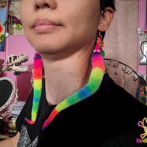 May include: A pair of novelty earrings in the shape of rainbow-coloured worms. Each earring has a fuzzy texture and is made up of a gradient of colours. The earrings are suspended from gold-coloured hooks and have googly eyes.