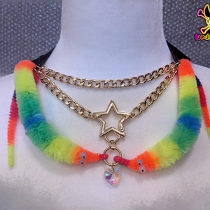 May include: A distinctive choker necklace with a rainbow-hued fuzzy worm design. It features gold-coloured chain accents, a star-shaped clasp, and a heart-shaped crystal pendant. The necklace is displayed on a white surface.