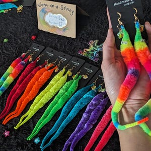 May include: Assortment of colorful fuzzy worm earrings. Each worm is made of yarn and has a gold hook. The earrings are displayed on a black surface with a sign that reads "Worm on a String" and "Earrings $20 Keychains $7".