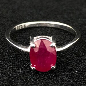 May include: A silver ring featuring a polished oval-cut deep red gemstone held by four prongs. The inner band of the ring is clearly stamped with the text S925, indicating the metal composition of the band. The ring has a thin, smooth silver band.