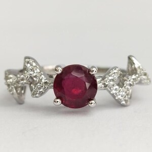 Op de afbeelding: Een zilveren ring met een ronde, dieprode robijn in het midden. De band heeft aan beide kanten een hoekig, grillig ontwerp, ingelegd met kleine, heldere, fonkelende witte steentjes die langs de metalen ringband doorlopen.