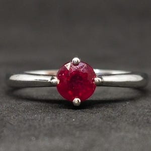 Puede incluir: Un anillo de tono plateado con una piedra preciosa única, de corte redondo y color rojo intenso, engastada en una montura de cuatro garras. El aro es liso y pulido, estrechándose ligeramente hacia la piedra central, situada en el centro.