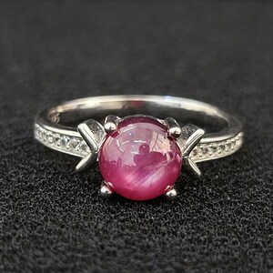 Può includere: Un anello color argento con una pietra preziosa centrale rotonda di colore rosa intenso, taglio cabochon con un sottile effetto gatteggiante. La fascia è decorata con piccole pietre chiare e scintillanti sui lati, fissate da quattro griffe.