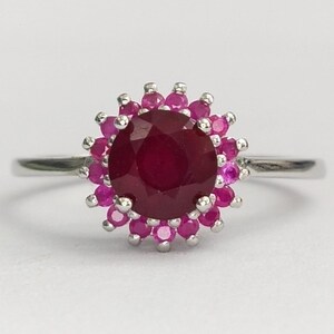 May include: A silver ring featuring a large, round, deep red centre stone surrounded by a halo of smaller, round, bright pink stones. The setting is metallic and polished, with a thin, smooth band extending from the central floral-shaped cluster.