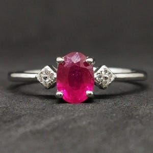 Può includere: Un anello color argento con una gemma rosso vivo dal taglio ovale tenuta da quattro griffe. Ai lati della pietra centrale si trova una piccola montatura quadrata contenente una singola pietra trasparente sfaccettata sulla fascia lucida.