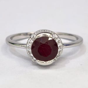 Könnte beinhalten: Ein silberfarbener Ring mit einem runden, tiefroten facettierten Edelstein in der Mitte. Der zentrale Stein ist von einem Kranz aus kleinen, klaren, funkelnden Steinen umgeben, die in eine polierte Metallschiene gefasst sind, die nach hinten schmaler wird.