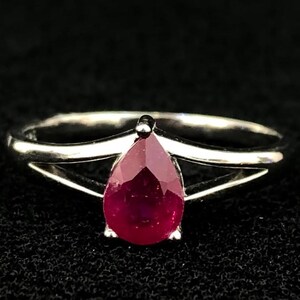 Puede incluir: Un anillo de tono plateado con una piedra preciosa de color rojo intenso en forma de pera, montada en un engaste de garras. La banda del anillo se divide en dos brazos metálicos finos y pulidos que se unen en la base de la piedra.
