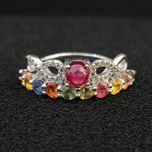 Può includere: Un anello color argento con un rubino cabochon rotondo centrale, affiancato da una fila di pietre preziose multicolori tra cui pietre blu, arancioni, verdi, gialle e rosa. La fascia presenta un intricato design traforato con piccoli accenti chiari.
