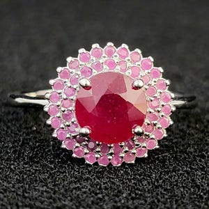 Könnte beinhalten: Ein silberfarbener Ring mit einem großen, runden, facettierten dunkelroten Mittelstein. Der Mittelstein ist von einem doppelten Halo aus kleinen, runden, hellrosa Steinen umgeben, die in einer gewellten Metallfassung sitzen.