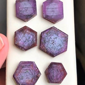 May include: Six hexagonal, faceted gemstones in a deep purple colour. The gems display a complex internal structure, with visible lines and patterns. The stones are arranged on a white surface, with a hand partially visible in the frame.