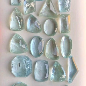 May include: A collection of 25 aquamarine gemstones in various shapes and sizes. The gemstones are faceted and have a light blue color.