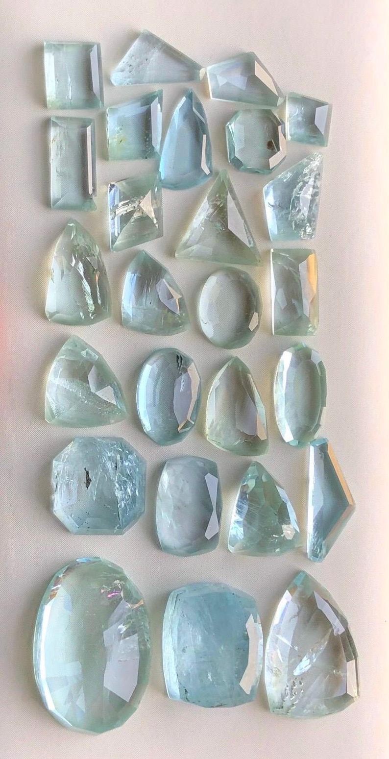 May include: A collection of 25 aquamarine gemstones in various shapes and sizes. The gemstones are faceted and have a light blue color.