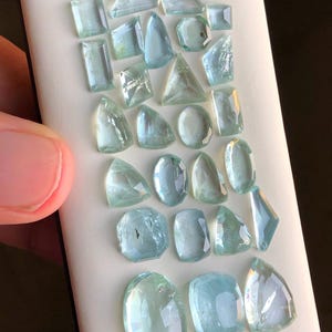 May include: A collection of 20 aquamarine gemstones of various shapes and sizes. The gemstones are arranged in rows on a white surface.