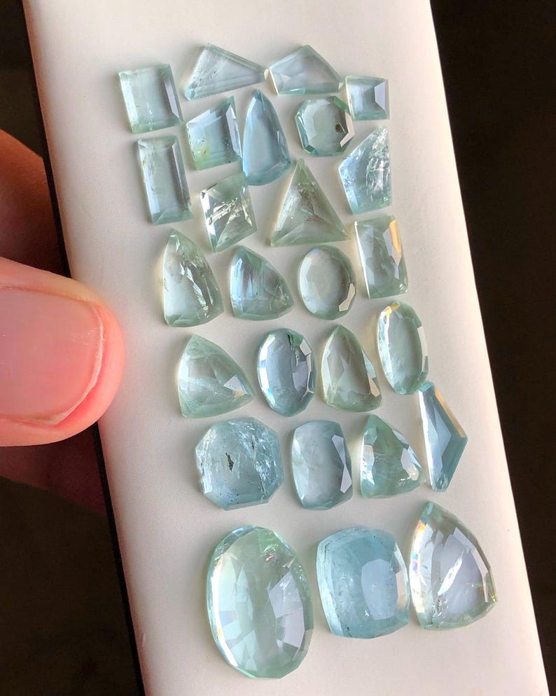 May include: A collection of 20 aquamarine gemstones of various shapes and sizes. The gemstones are arranged in rows on a white surface.