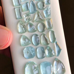 May include: A collection of 22 aquamarine gemstones of various shapes and sizes. The gemstones are arranged in rows on a white surface.