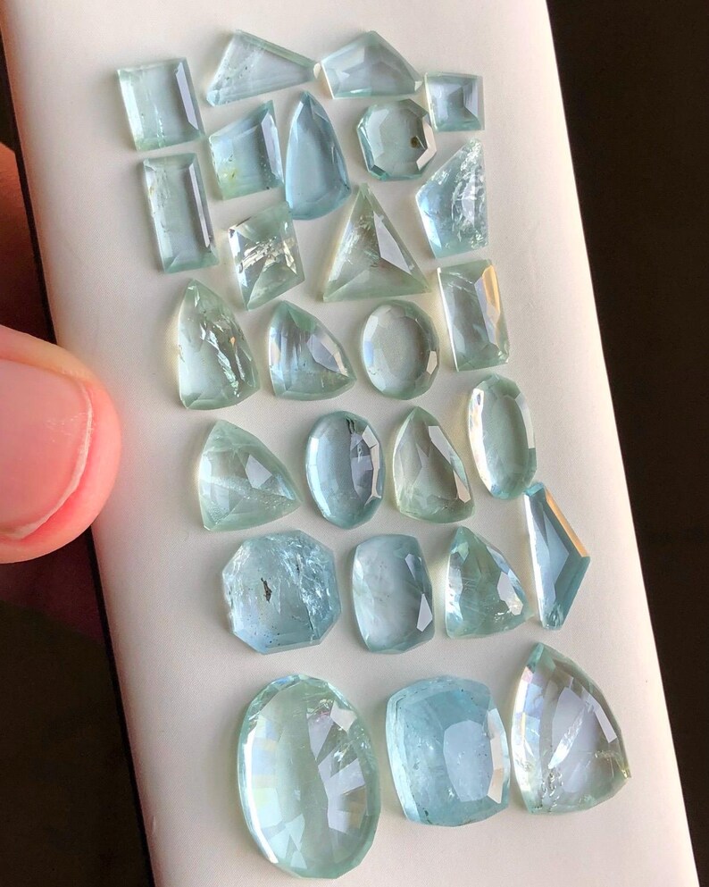 May include: A collection of 22 aquamarine gemstones of various shapes and sizes. The gemstones are arranged in rows on a white surface.
