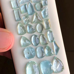 May include: A collection of 20 aquamarine gemstones of various shapes and sizes. The gemstones are arranged on a white surface.