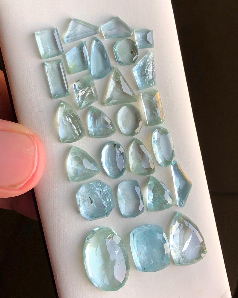 May include: A collection of 20 aquamarine gemstones of various shapes and sizes. The gemstones are arranged on a white surface.