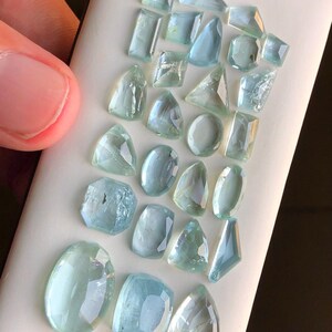 May include: A collection of aquamarine gemstones of various shapes and sizes, displayed on a white surface.