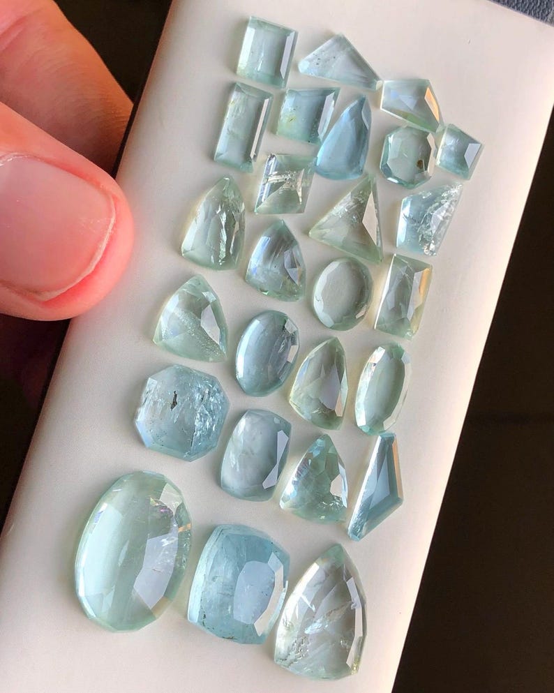 May include: A collection of aquamarine gemstones of various shapes and sizes, displayed on a white surface.