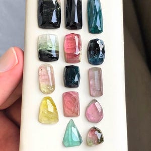 May include: Assortment of faceted tourmaline gemstones in various colors, including black, green, pink, and yellow. The gemstones are displayed on a white surface, showcasing their clarity and unique shapes. A hand is partially visible in the image.