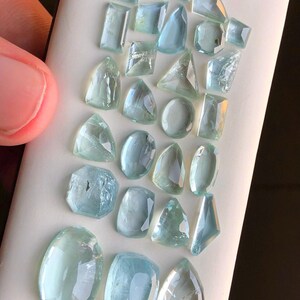 May include: A collection of aquamarine gemstones in various shapes and sizes, including oval, round, and rectangular cuts. The stones are a light blue color and have a translucent appearance.