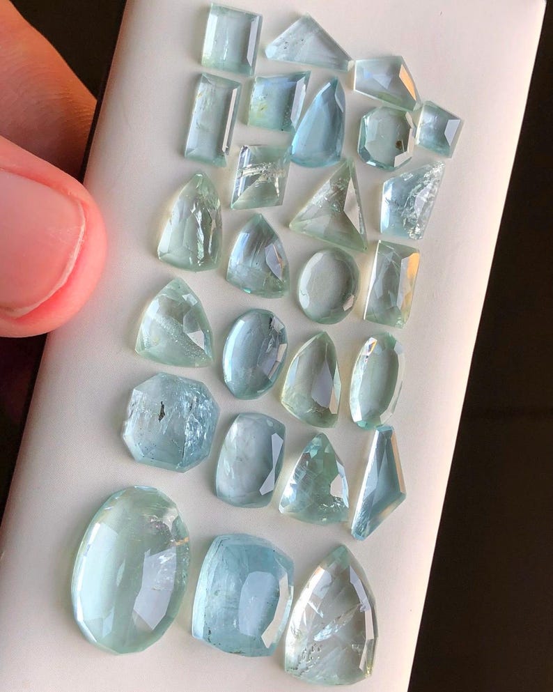 May include: A collection of aquamarine gemstones in various shapes and sizes, including oval, round, and rectangular cuts. The stones are a light blue color and have a translucent appearance.