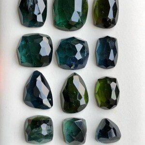 May include: A collection of twelve faceted gemstones in shades of green and teal. The gemstones vary in shape, including square, oval, and triangular cuts. The gems are displayed on a white surface, showcasing their polished surfaces and internal clarity.