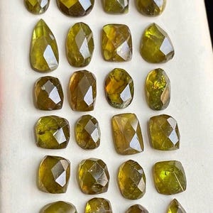 May include: Twenty-one faceted gemstones in diverse shapes, such as oval, rectangular, and teardrop. The gems exhibit olive green and golden-brown shades, with multiple facets reflecting light. The gemstones are arranged on a white surface.