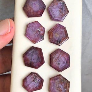 May include: Eight hexagonal gemstones in a deep red-purple colour. The gems have a faceted design, with visible internal structures and colour variations. The stones are arranged on a light-coloured surface, showcasing their polished appearance.
