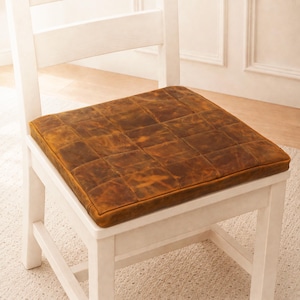 Puede incluir: Un coj&iacute;n de asiento cuadrado con un dise&ntilde;o de parches de cuadrados de cuero marr&oacute;n desgastado. El coj&iacute;n est&aacute; colocado sobre una silla de madera blanca. El cuero tiene una textura variada y moteada con tonos marrones claros y oscuros.