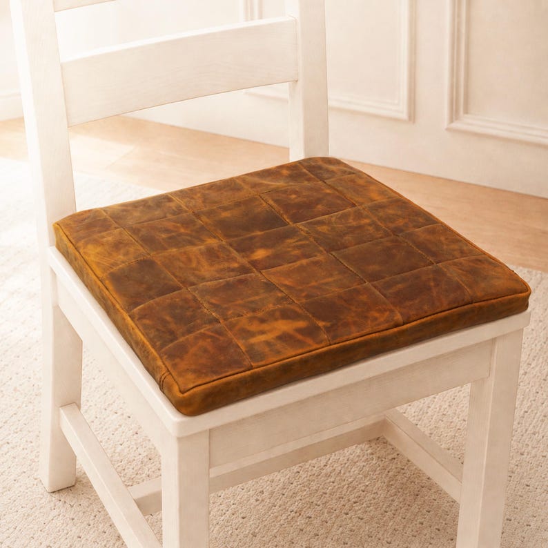 Puede incluir: Un coj&iacute;n de asiento cuadrado con un dise&ntilde;o de parches de cuadrados de cuero marr&oacute;n desgastado. El coj&iacute;n est&aacute; colocado sobre una silla de madera blanca. El cuero tiene una textura variada y moteada con tonos marrones claros y oscuros.
