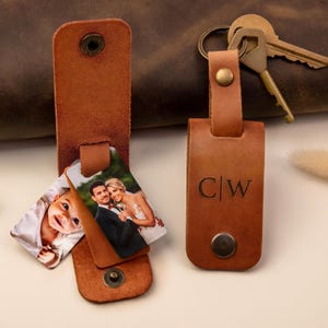 May include: Two tan leather key organizers with metal snap buttons. One organizer is open, revealing two rectangular photo inserts showing a baby and a couple. The other organizer is closed and features the embossed letters C|W with a metal key ring.
