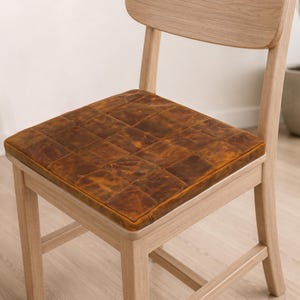 Puede incluir: Una silla de madera clara con un coj&iacute;n de asiento cuadrado hecho de cuero marr&oacute;n con un patr&oacute;n de cuadr&iacute;cula visible. El coj&iacute;n tiene un acabado texturizado y desgastado, y est&aacute; ajustado con precisi&oacute;n sobre el marco de madera de la silla.