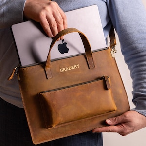 May include: A brown leather laptop bag with a handle and a detachable shoulder strap. The bag has a front pocket with a zipper and the name "BRADLEY" is embossed on the front. A silver laptop is inside the bag.