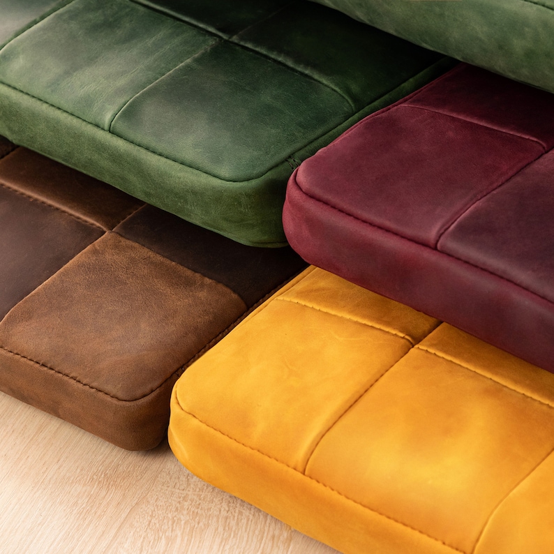 Puede incluir: Una pila de cuatro estuches rectangulares de cuero en verde, burdeos, marr&oacute;n y amarillo. Cada estuche presenta un acabado mate liso, esquinas redondeadas y costuras visibles que crean un patr&oacute;n de cuadr&iacute;cula sobre la superficie del cuero.