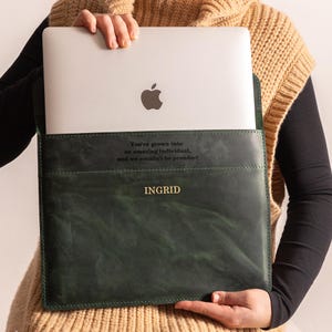 May include: A dark green leather laptop sleeve with the name "INGRID" in gold lettering. The sleeve holds a silver laptop with an Apple logo. Text on the sleeve reads "You've grown into an amazing individual, and we couldn't be prouder!"