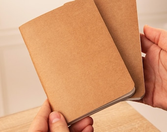 May include: Two small notebooks with plain, light brown kraft paper covers. The notebooks have rounded corners and a simple, unadorned design. The covers are made of a textured, matte paper material, and the pages inside are visible along the edges.