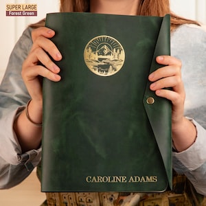 May include: A forest green leather journal with a gold embossed mountain scene and the name "CAROLINE ADAMS" on the front.