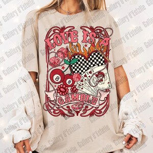 May include: Beige t-shirt with a retro graphic design. The design features the words "LOVE IS A GAMBLE" in pink, along with dice, playing cards, cherries, and a checkered heart. The shirt has short sleeves.