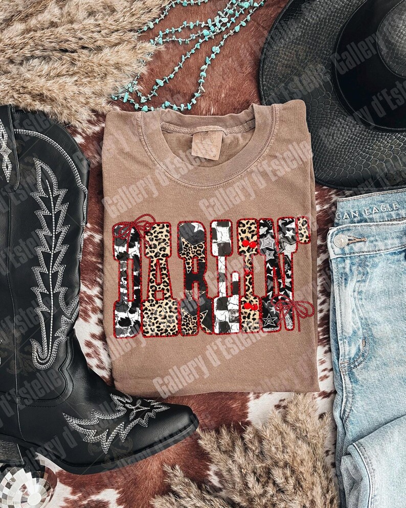 May include: A tan t-shirt with the word "DARLIN" in a decorative font with animal print and star accents. The shirt is surrounded by a black cowboy boot, a black hat, and blue jeans.
