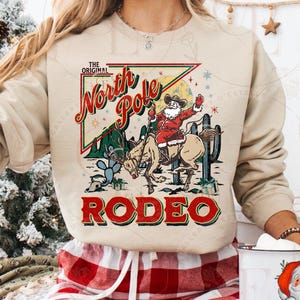 May include: Beige sweatshirt with a graphic of Santa Claus riding a horse in a rodeo scene. The text on the sweatshirt reads "The Original North Pole Rodeo" in red and green. The design includes cacti and stars.