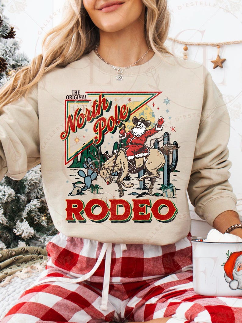 May include: Beige sweatshirt with a graphic of Santa Claus riding a horse in a rodeo scene. The text on the sweatshirt reads "The Original North Pole Rodeo" in red and green. The design includes cacti and stars.