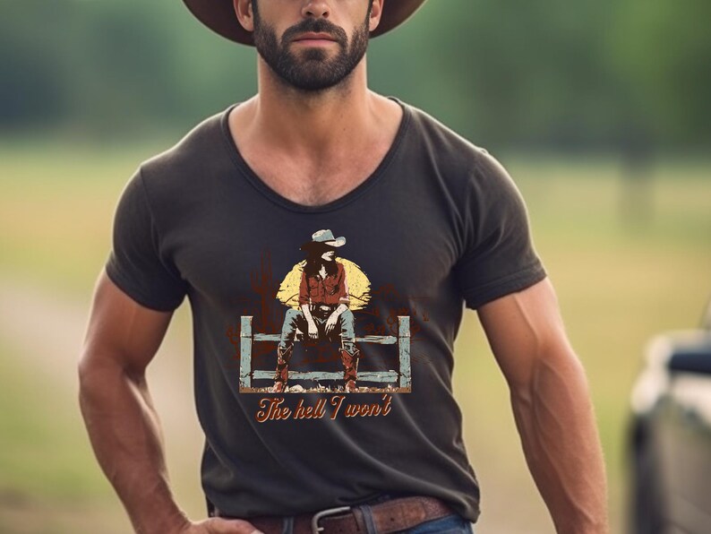 Op de afbeelding: Een donkerbruin V-hals T-shirt met een vintage afbeelding van een vrouw die een cowboyhoed draagt en op een hek zit. De tekst "The hell I won't" is onder de afbeelding gedrukt.