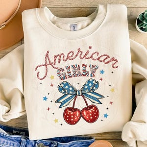 May include: A white sweatshirt with the text "American Girl" in red and blue lettering. The text is surrounded by stars and a red, white and blue bow with cherries hanging from it.