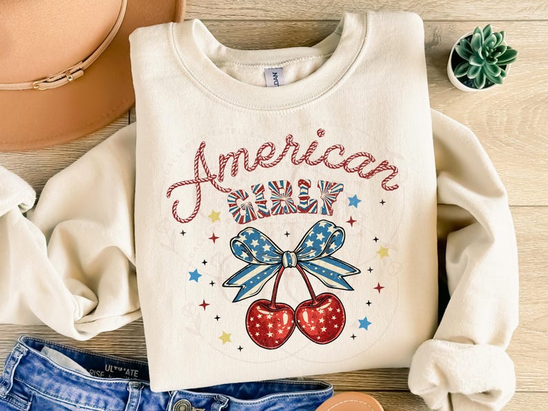 May include: A white sweatshirt with the text "American Girl" in red and blue lettering. The text is surrounded by stars and a red, white and blue bow with cherries hanging from it.