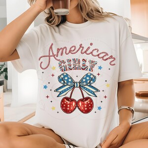 May include: White t-shirt featuring a red, white and blue cherry graphic and the text "American Girl" in a rope-like font.