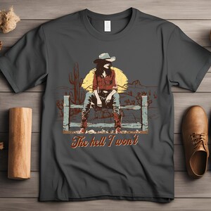 Op de afbeelding: Een donkergrijs T-shirt met een vintage illustratie van een vrouw die een cowboyhoed en laarzen draagt ​​en op een hek zit. De tekst "The hell I won't" is onder de illustratie gedrukt.